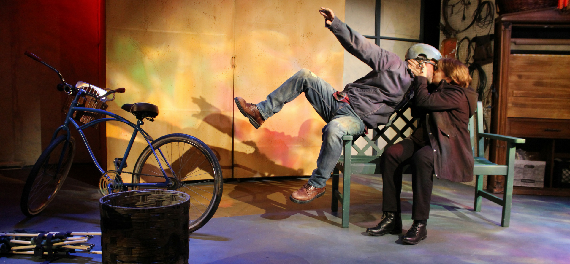 Man and woman on stage in play kissing; the man had just fallen off of a bike that is in the background